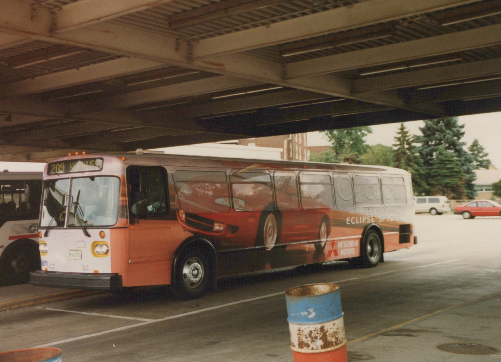 A Pace bus with fleet number 8871 is parked under an elevated structure, featuring a large advertisement for the Mitsubishi Eclipse Spyder on its side. The bus shows route 209 Northwest on its destination sign.