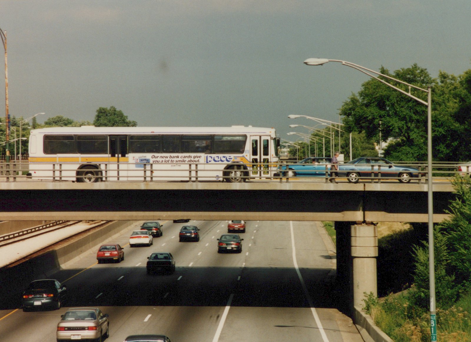 A white Pace bus with a yellow stripe drives across a concrete bridge over a multi-lane highway with cars below and a pedestrian walking beside it.
