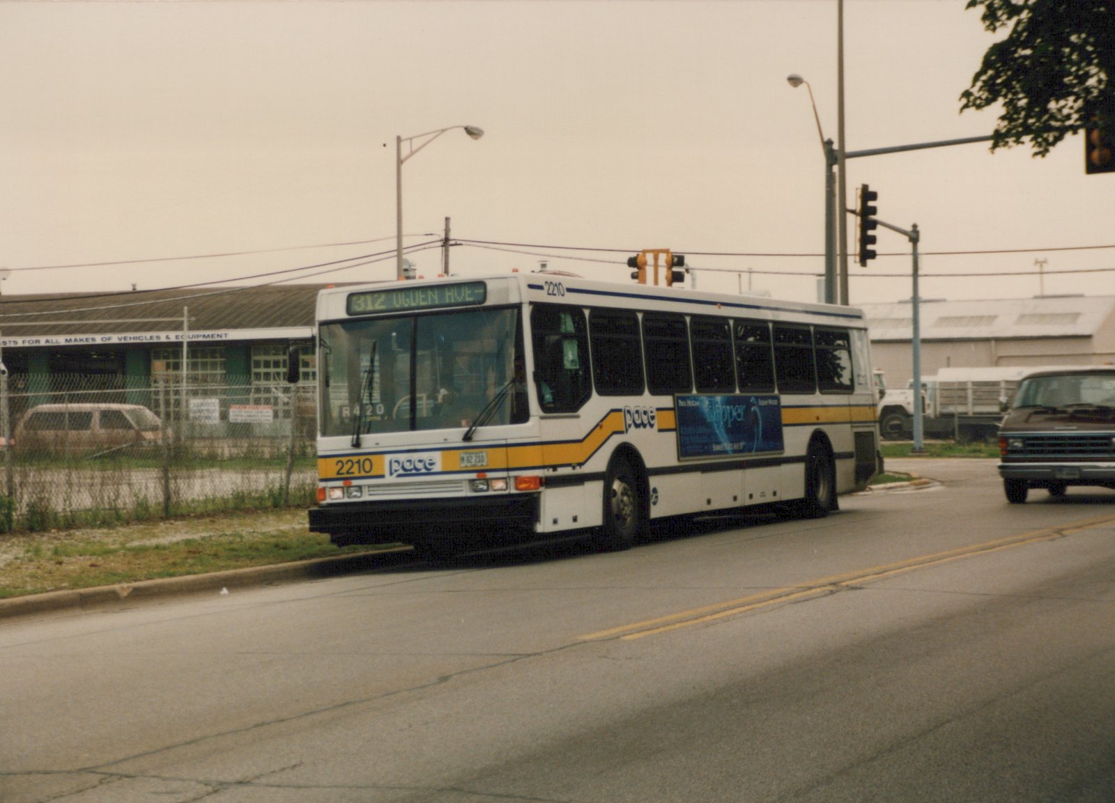 A white Pace bus with blue and yellow stripes, numbered 2210, is shown from a three-quarter front view on a street, with a destination sign reading '312 OGDEN AVE'.