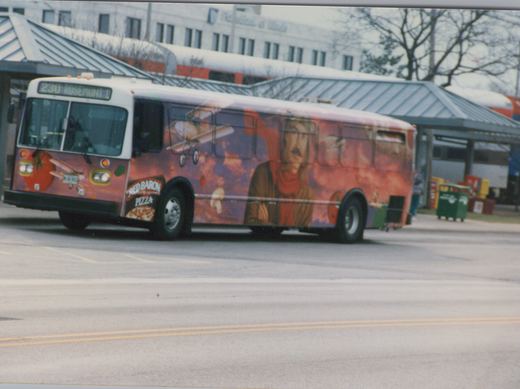 A white and red bus with a full Red Baron Pizza ad, showing route 230 to Rosemont, is on a street in front of a train station with a double-decker Amtrak train behind it.