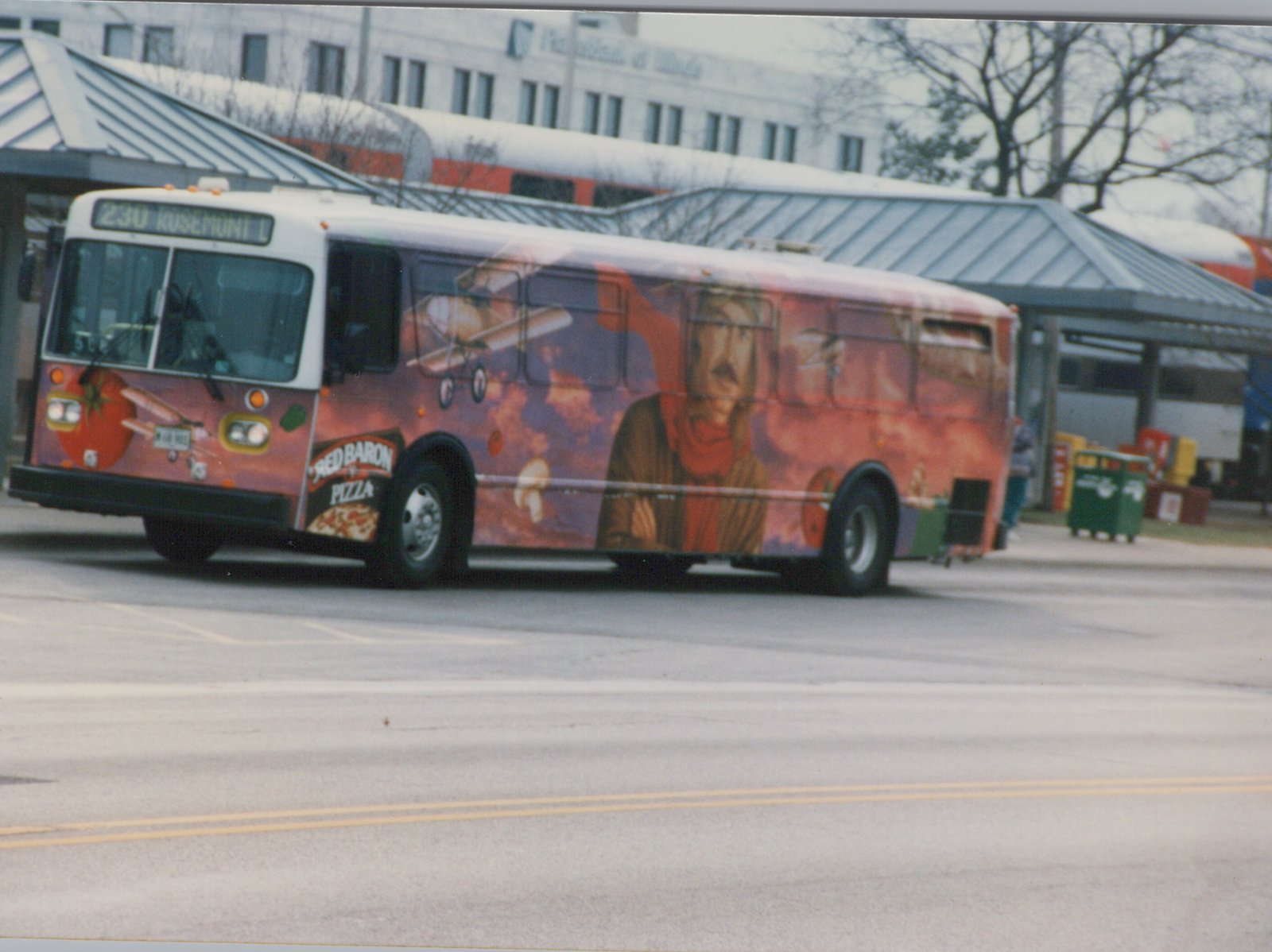 A white and red bus with a full Red Baron Pizza ad, showing route 230 to Rosemont, is on a street in front of a train station with a double-decker Amtrak train behind it.
