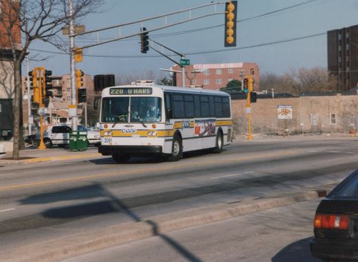 A white Pace bus with yellow and blue stripes, marked 2091 and heading to O'Hare, is driving down a street with bare trees and traffic lights under a clear blue sky.
