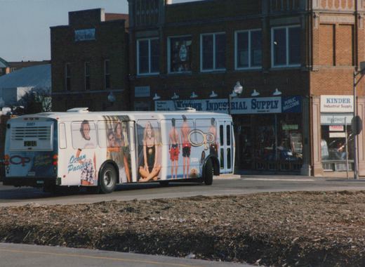 A white Pace bus with an Ocean Pacific advertisement is parked on a city street in front of a store named Kinder Industrial Supply.