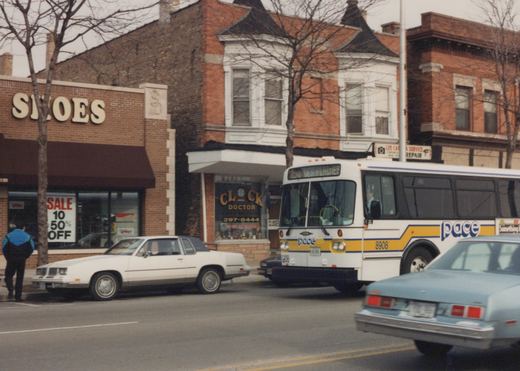 A white and yellow Pace bus is driving down a street in Des Plaines, Illinois, past brick storefronts and parked cars on a cloudy winter day.