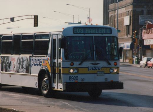 A white Pace motor bus with blue and yellow stripes, numbered 8920, is shown from a three-quarter front view on a city street, displaying route 209 Harlem on its front sign.