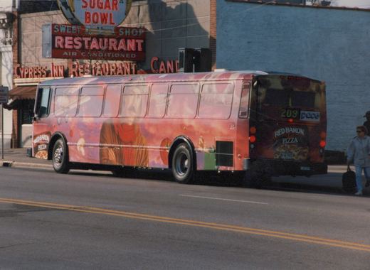 A red and orange bus with a full Red Baron Pizza ad, showing the number 203 and Pace logo on the back, parked on a street beside a building with signs for Sugar Bowl and Cypress Inn Restaurant.