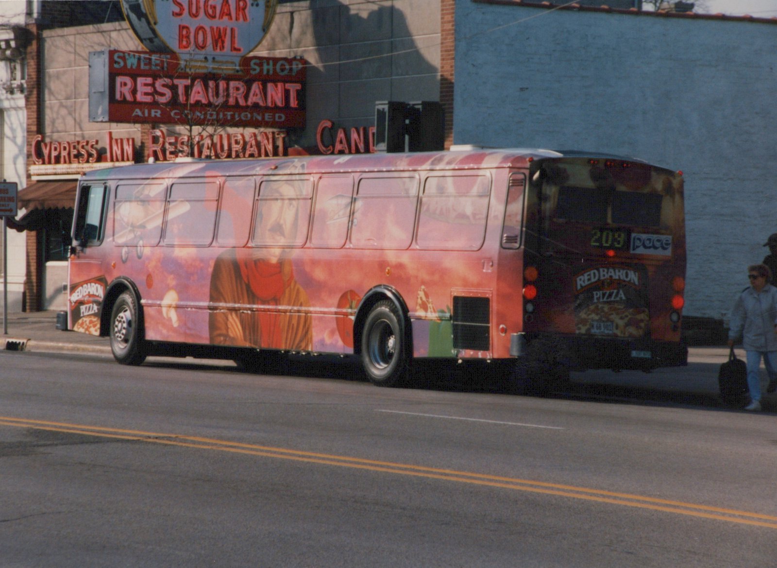 A red and orange bus with a full Red Baron Pizza ad, showing the number 203 and Pace logo on the back, parked on a street beside a building with signs for Sugar Bowl and Cypress Inn Restaurant.