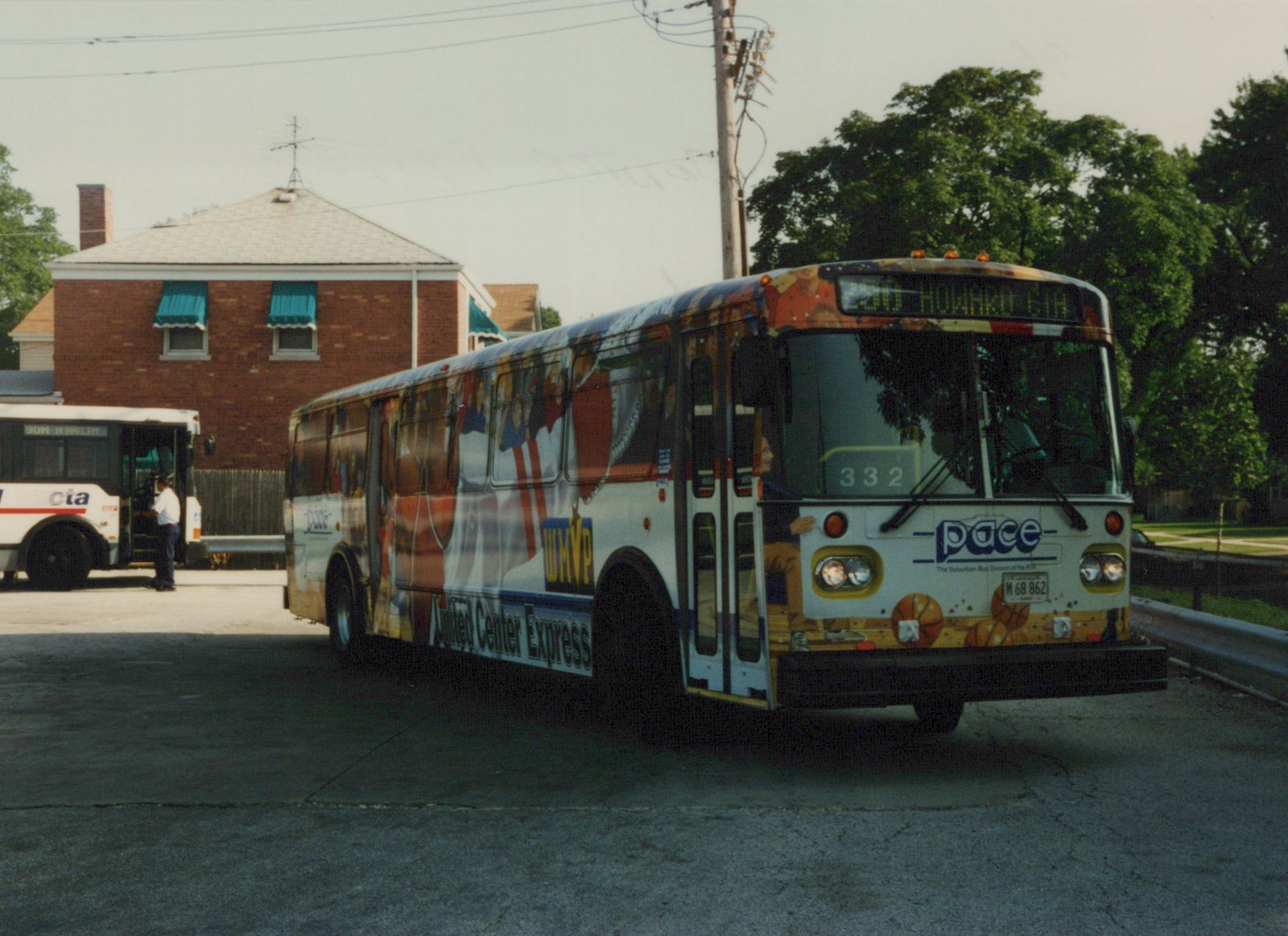 A white Pace bus with a colorful basketball-themed wrap is parked on a street, with a CTA bus visible in the background. The Pace bus displays the number 332 and route 330 Howard Ctr.