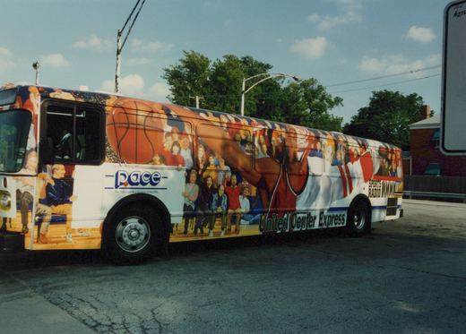A side view of a white Pace bus with a colorful wraparound advertisement featuring a basketball player and spectators, parked on a paved street under a partly cloudy sky.