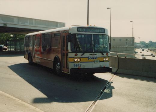A white and maroon Pace bus, number 8852, is parked on a concrete median next to a highway; its front sign reads '209 NORTHWEST' and it is beneath a bridge marked '209'.