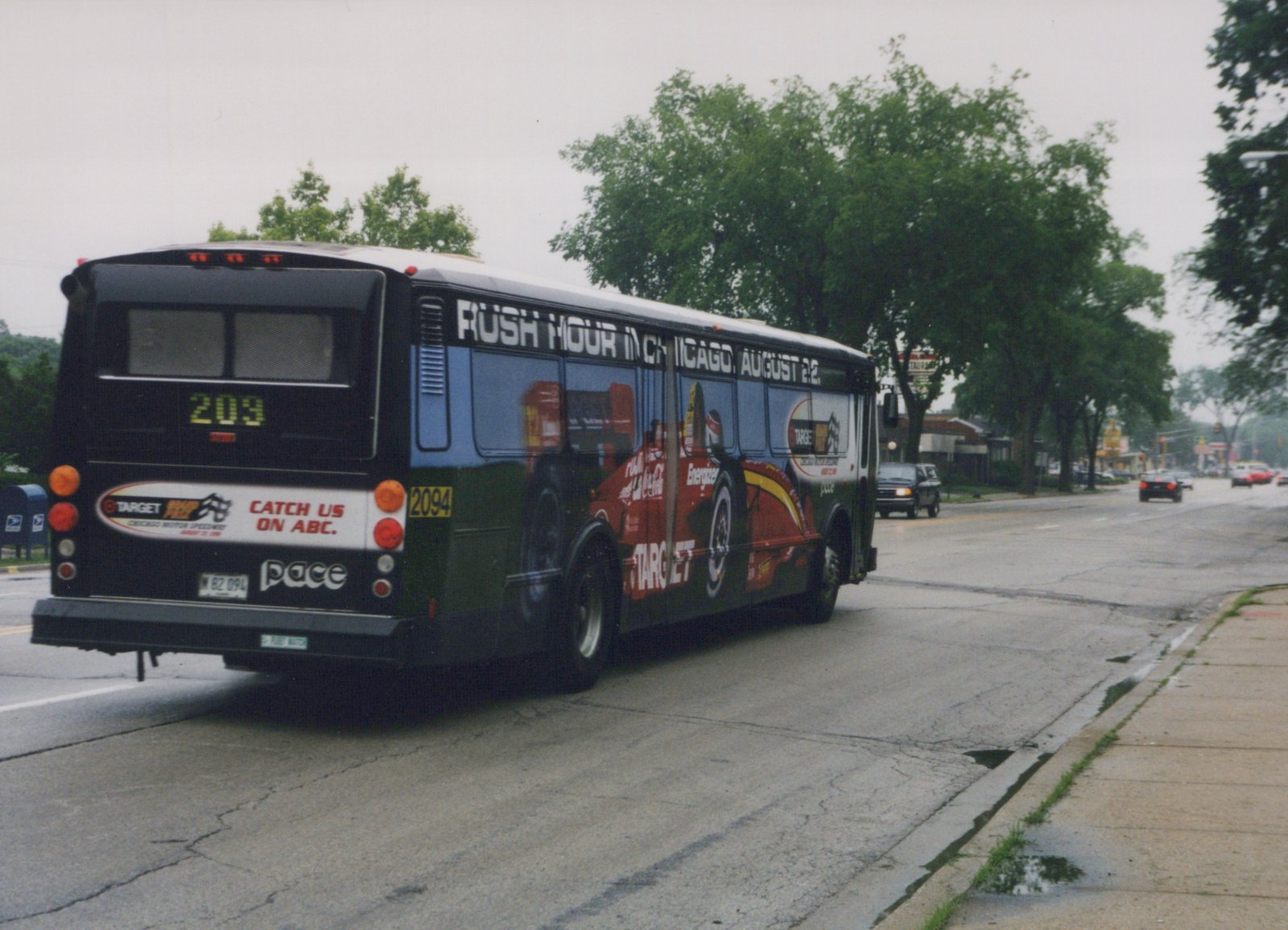 Three-quarter rear view of a black Pace motor bus with a red and white 'Rush Hour' movie advertisement, driving on a wet street with trees and other cars in the background.