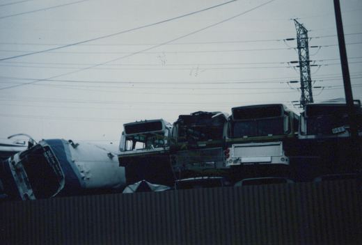 Rear view of several decommissioned buses in a scrap yard, with the closest one showing fleet number 9681, under a gray, overcast sky with power lines overhead.