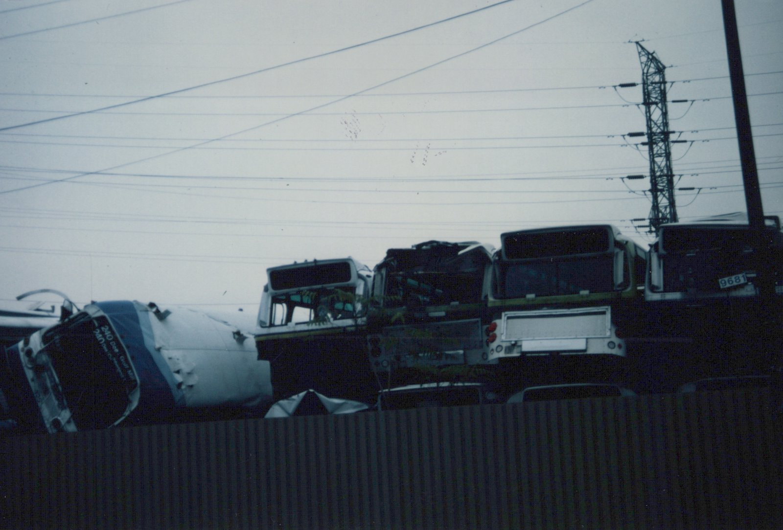 Rear view of several decommissioned buses in a scrap yard, with the closest one showing fleet number 9681, under a gray, overcast sky with power lines overhead.