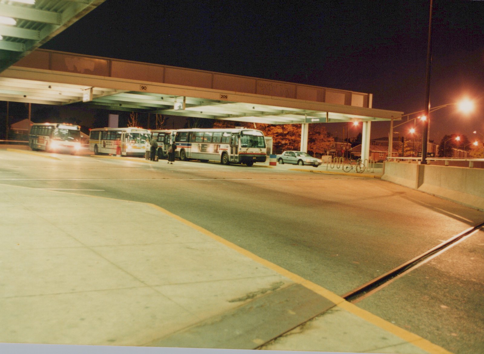 A nighttime scene of several Pace motor buses stopped at a terminal located in an expressway median, with people waiting, under a lighted shelter with route numbers 90 and 209 visible, and a car and bicycle parked nearby.