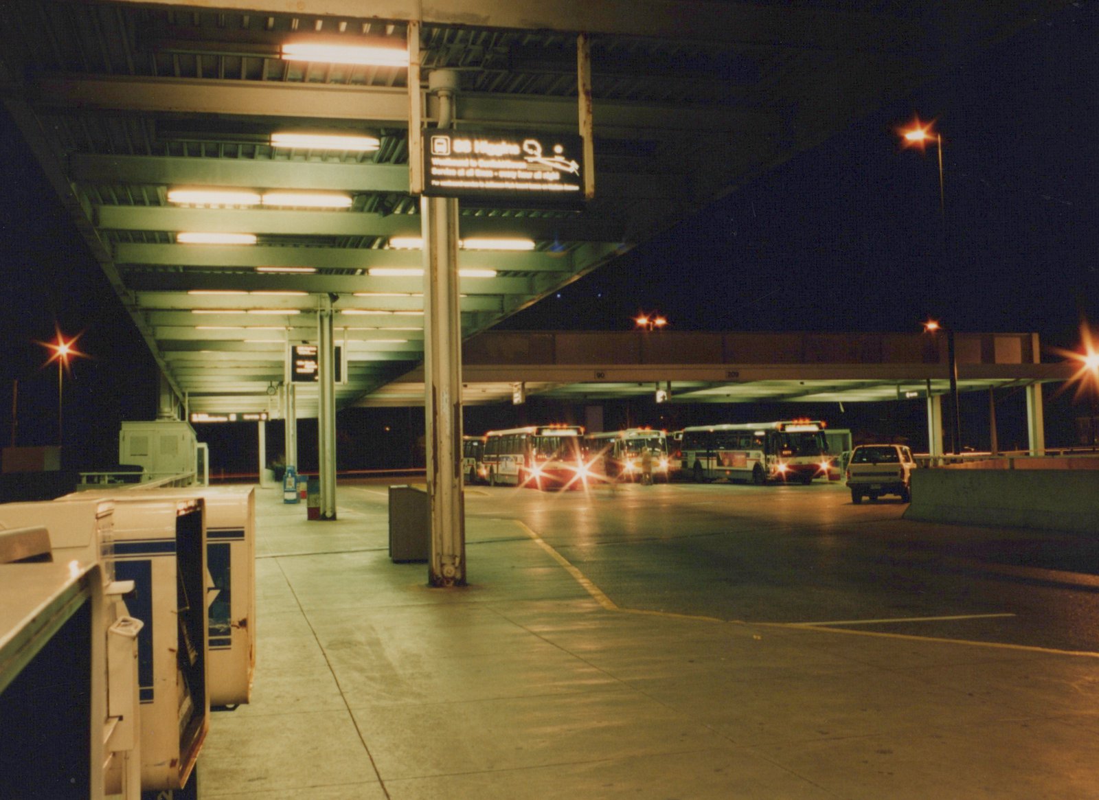 A nighttime view of a bus terminal with lit overhead signs, several parked buses, and a pickup truck in the background.