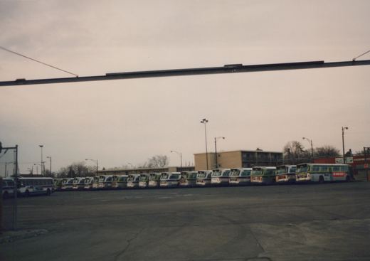 A wide shot of a bus garage with a row of CTA buses parked in an empty lot, under an overpass, with buildings and leafless trees in the background on a cloudy day.