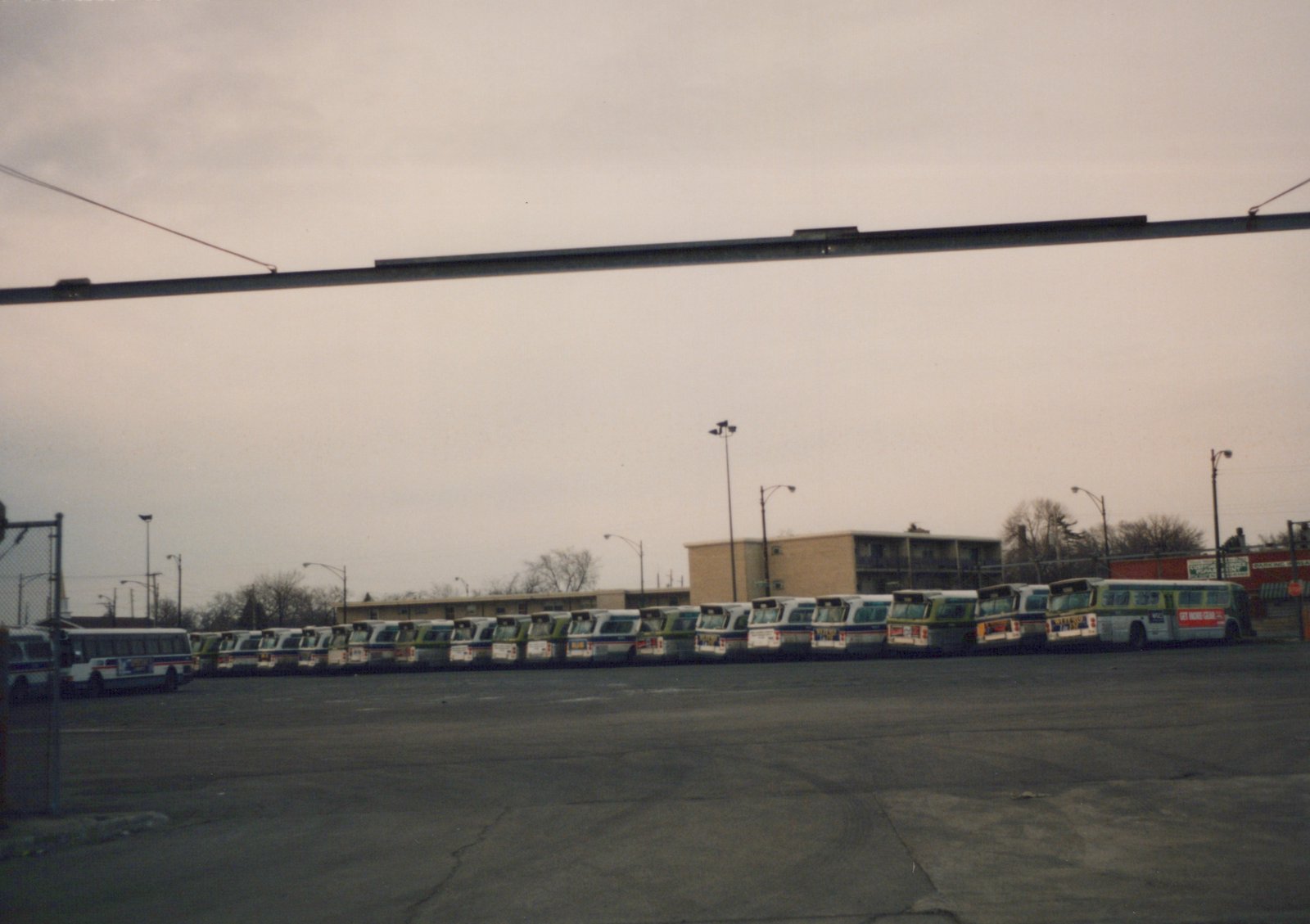 A wide shot of a bus garage with a row of CTA buses parked in an empty lot, under an overpass, with buildings and leafless trees in the background on a cloudy day.