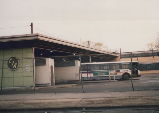 A side view of a white CTA motor bus parked next to a green-tiled garage building under an overcast sky.