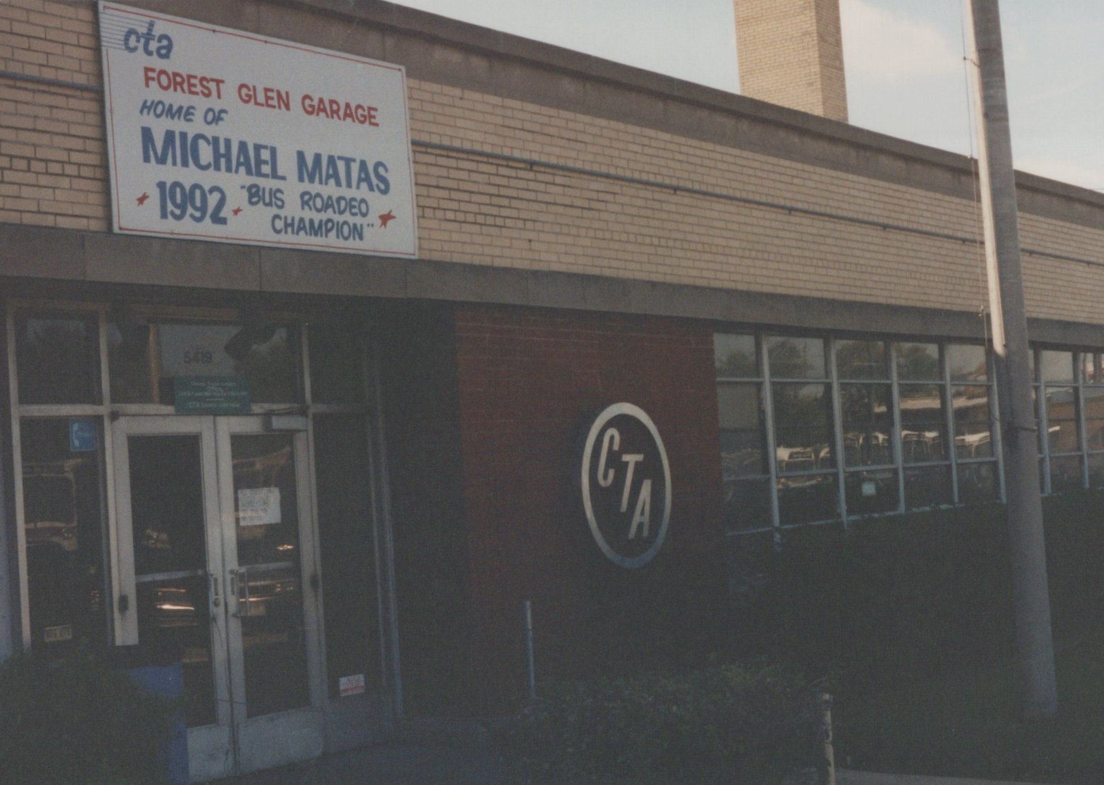 A brick building with a large sign reading 'CTA Forest Glen Garage, Home of Michael Matas, 1992 Bus Roadeo Champion', featuring the CTA logo and a view into the garage through large windows.