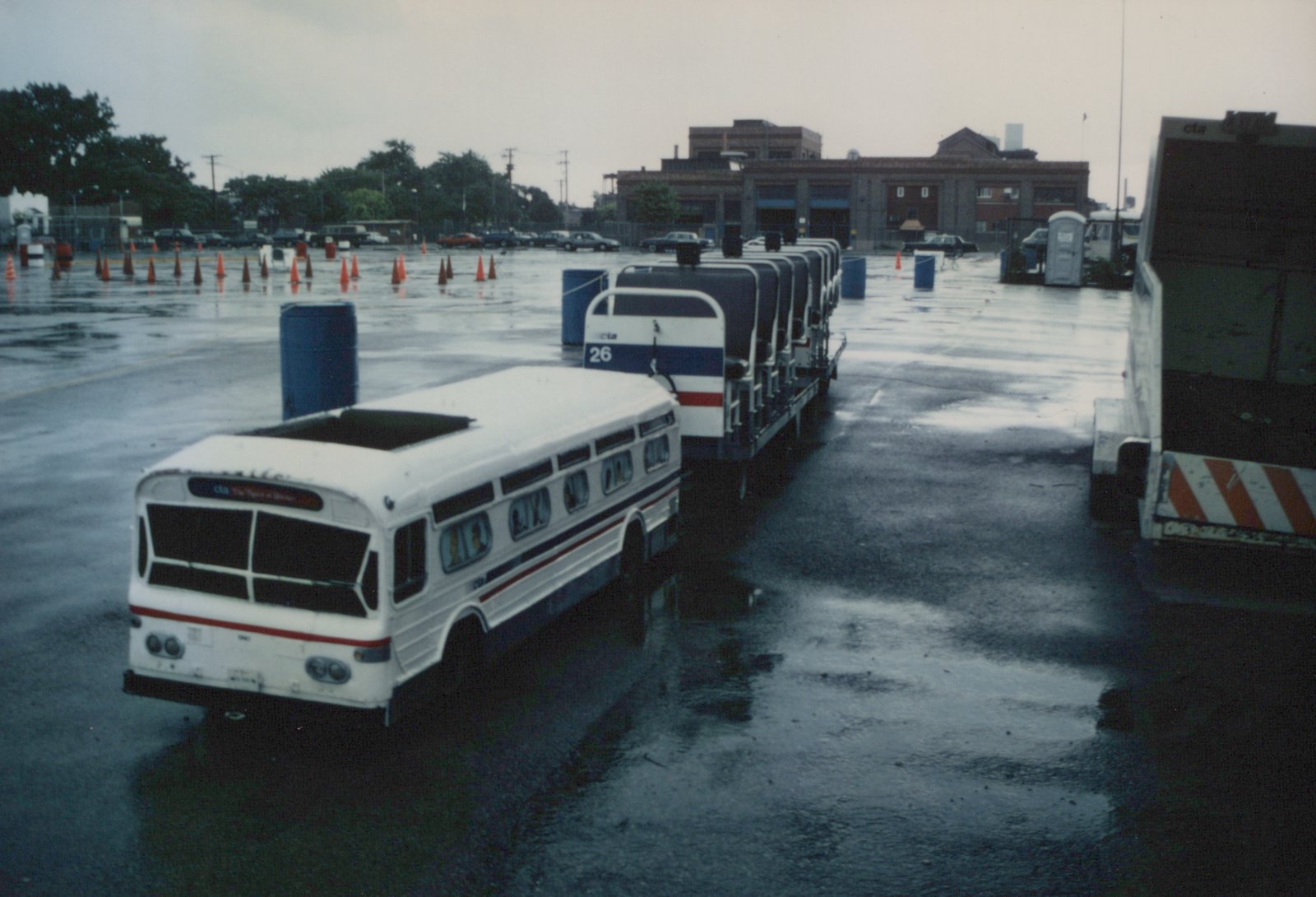 A white CTA bus with blue and red stripes, number 26, parked on wet pavement in a bus yard with orange cones and other vehicles in the background.