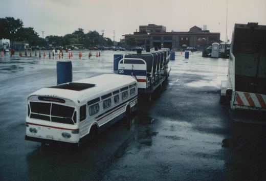 A white CTA bus with blue and red stripes, number 26, parked on wet pavement in a bus yard with orange cones and other vehicles in the background.