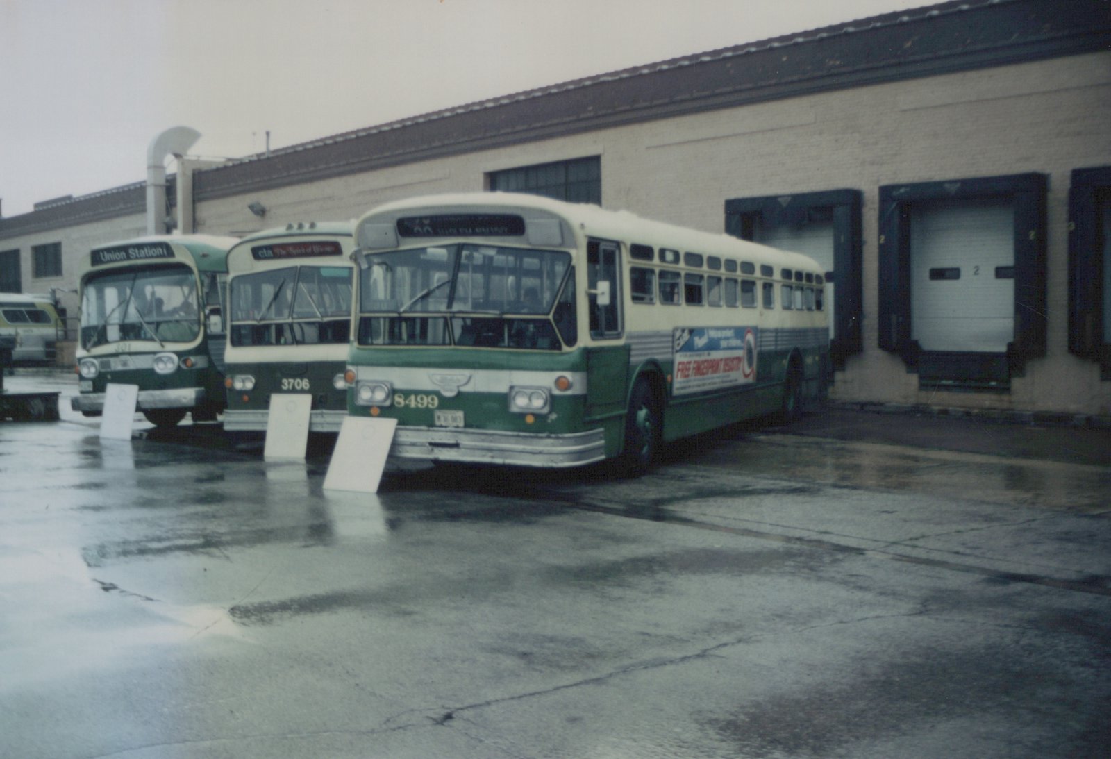 Three green and cream CTA buses parked side-by-side in front of a garage with large roll-up doors, the ground is wet and reflective.