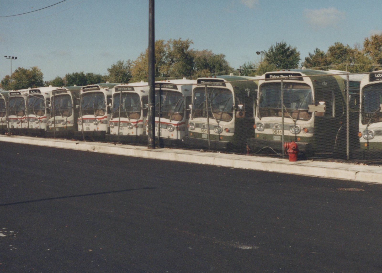 A row of green and cream Chicago Transit Authority buses parked behind a chain-link fence on an asphalt lot under a blue sky with trees in the background.