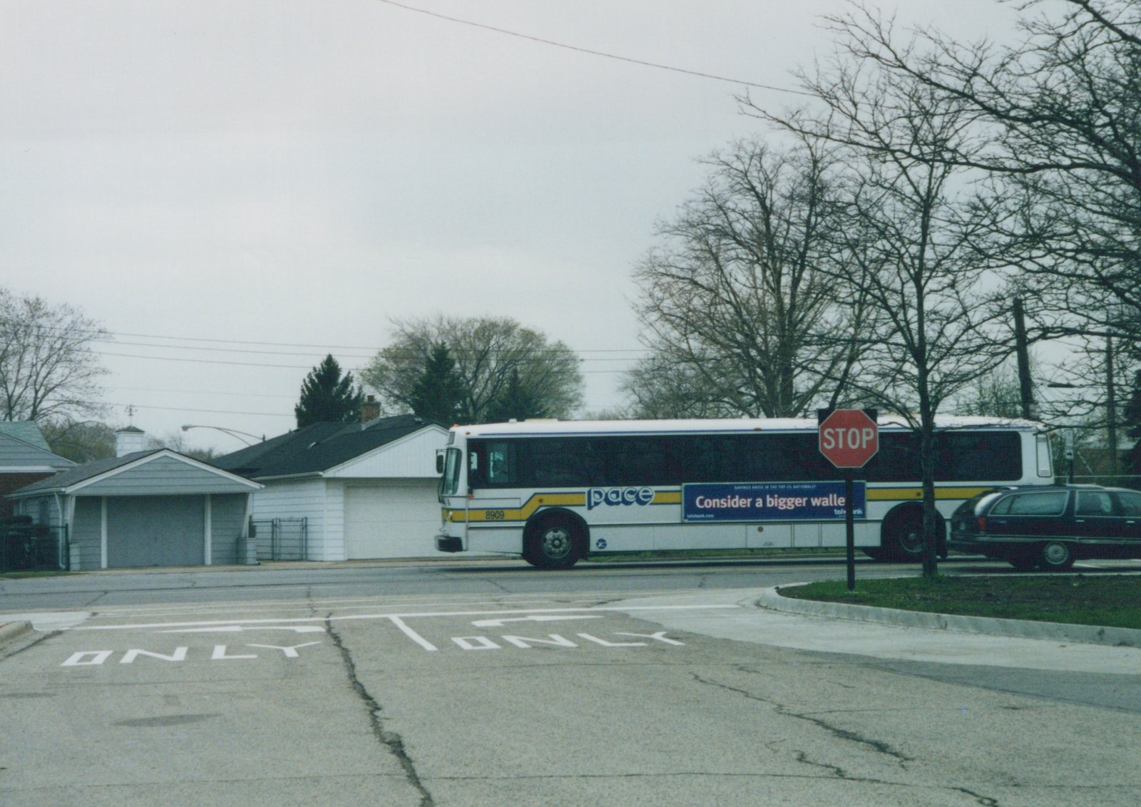 A white Pace bus with a yellow stripe, number 8909, is parked on a street next to a stop sign and a parked station wagon, with bare trees and houses in the background.
