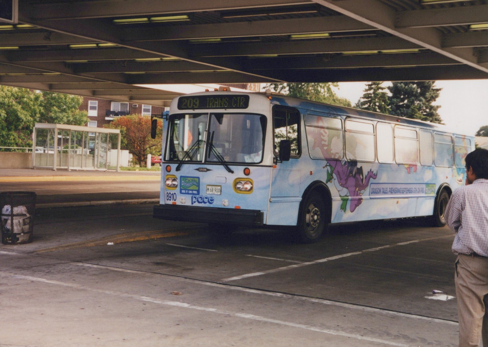 A light blue Pace bus with a cartoon dragon on its side is parked under a concrete overpass, with one passenger standing beside it.