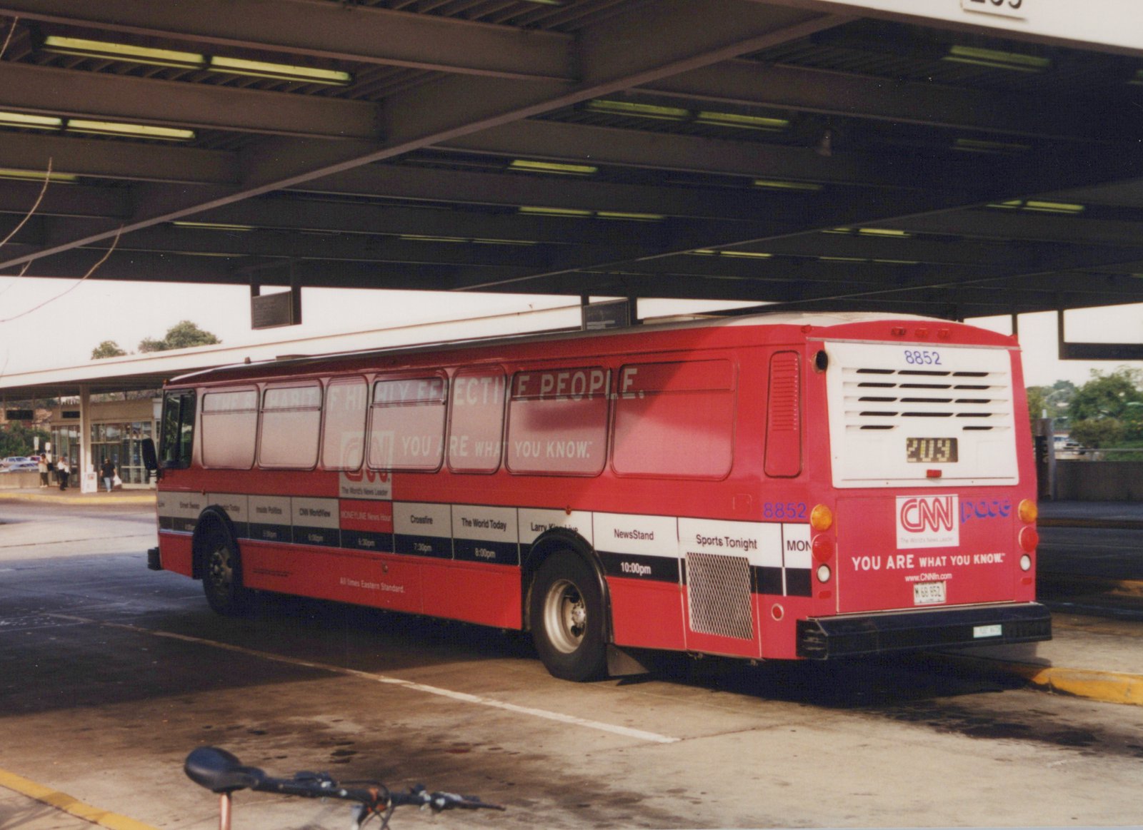 A red Pace bus with the number 8852 and a CNN ad is parked under a shelter, with people nearby and a bicycle in the foreground.