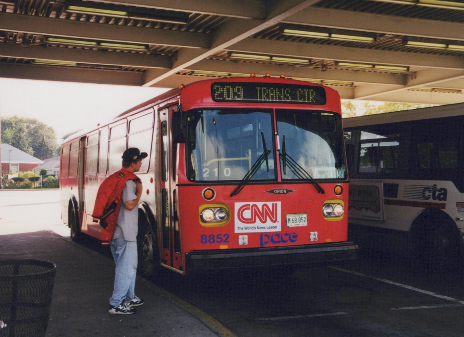 A red and white Pace bus, number 8852, with a '209 TRANS CTR' destination sign, is parked under a shelter with a person standing beside it.