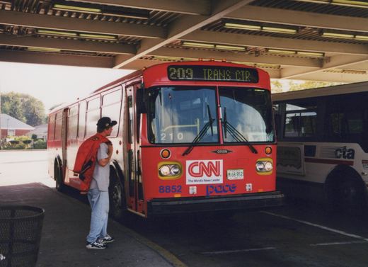 A red and white Pace bus, number 8852, with a '209 TRANS CTR' destination sign, is parked under a shelter with a person standing beside it.
