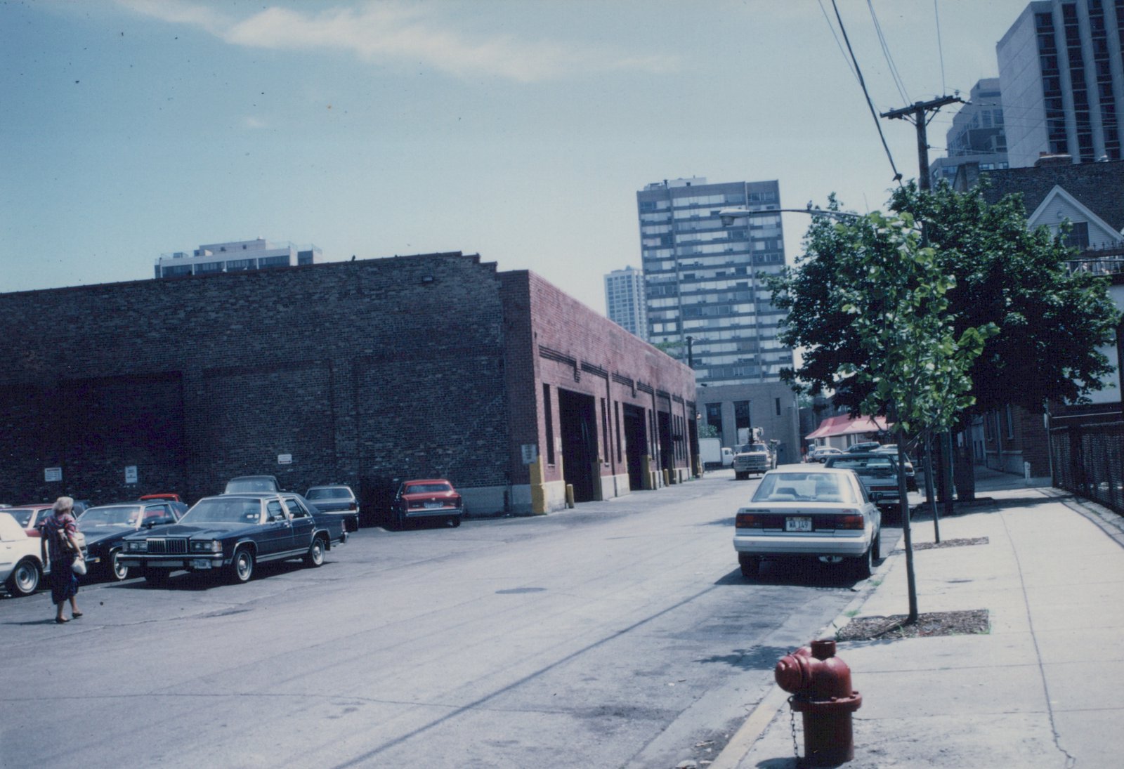 A street view in Chicago showing several parked cars, including a white sedan with license plate MA 149, a pedestrian walking, a large brick building, and high-rise apartments in the distance.