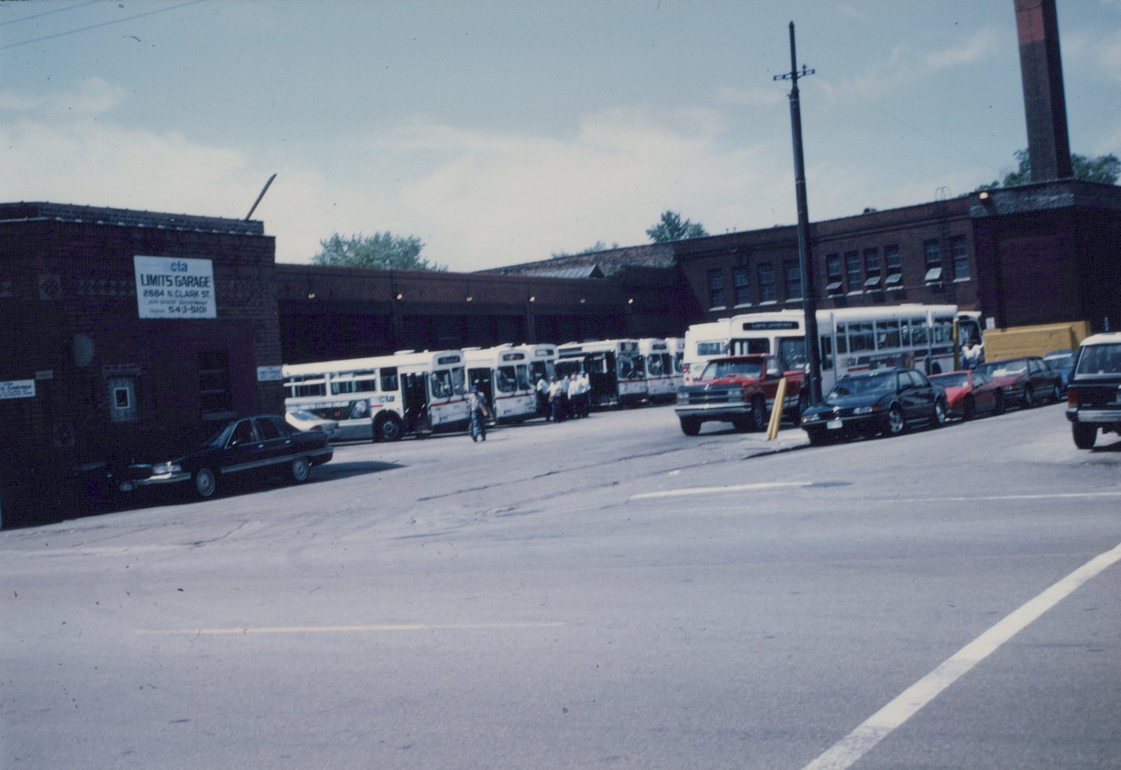 A lineup of white CTA buses with red and blue stripes parked in front of a brick building labeled 'cta LIMITS GARAGE' with several cars and people visible in the scene.
