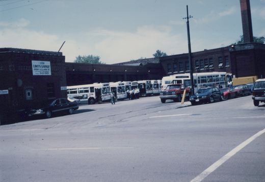 A lineup of white CTA buses with red and blue stripes parked in front of a brick building labeled 'cta LIMITS GARAGE' with several cars and people visible in the scene.