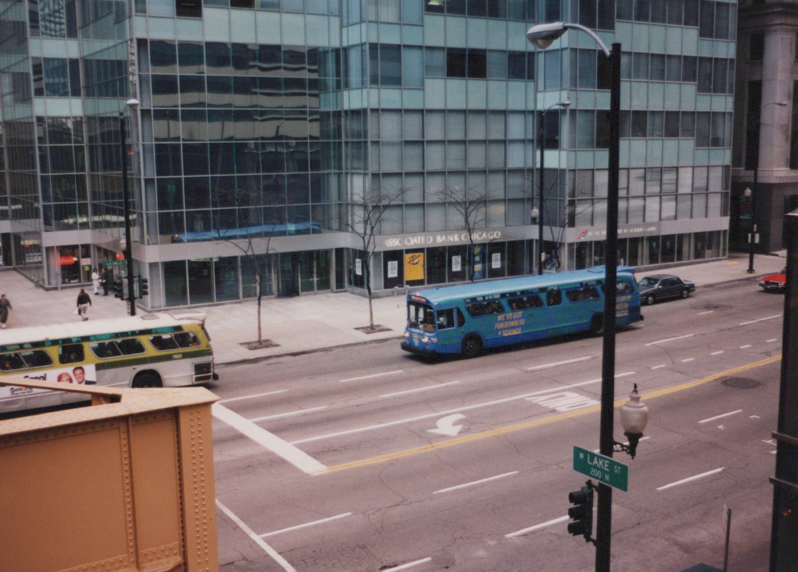 Two Chicago Transit Authority buses on a city street. One is blue with a science-themed ad, the other is white with a green stripe. A green street sign reads 'W LAKE ST' and a large glass building stands behind them.