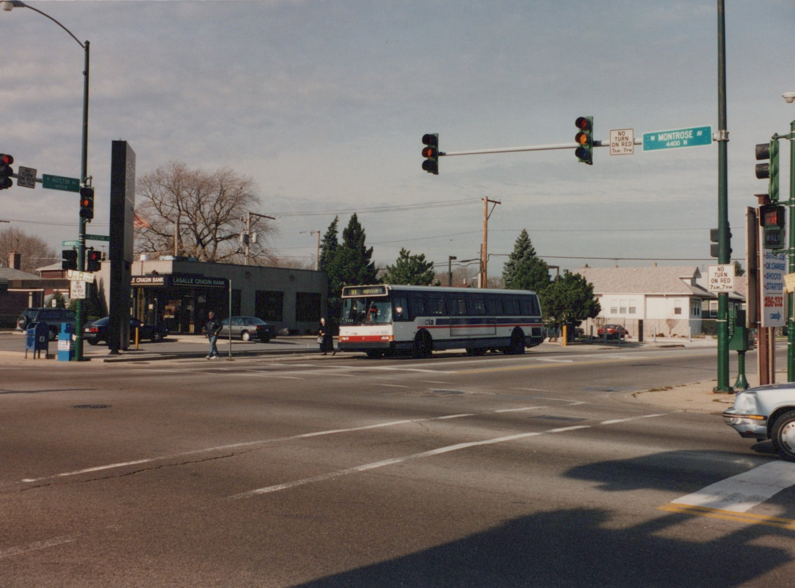 A white CTA bus with red and blue stripes, number 9134, is stopped at a street intersection with traffic lights and pedestrians nearby. The street signs read N Austin Ave and W Montrose Ave.