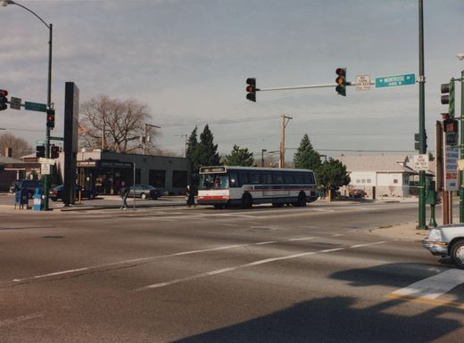 A white CTA bus with red and blue stripes, number 9134, is stopped at a street intersection with traffic lights and pedestrians nearby. The street signs read N Austin Ave and W Montrose Ave.