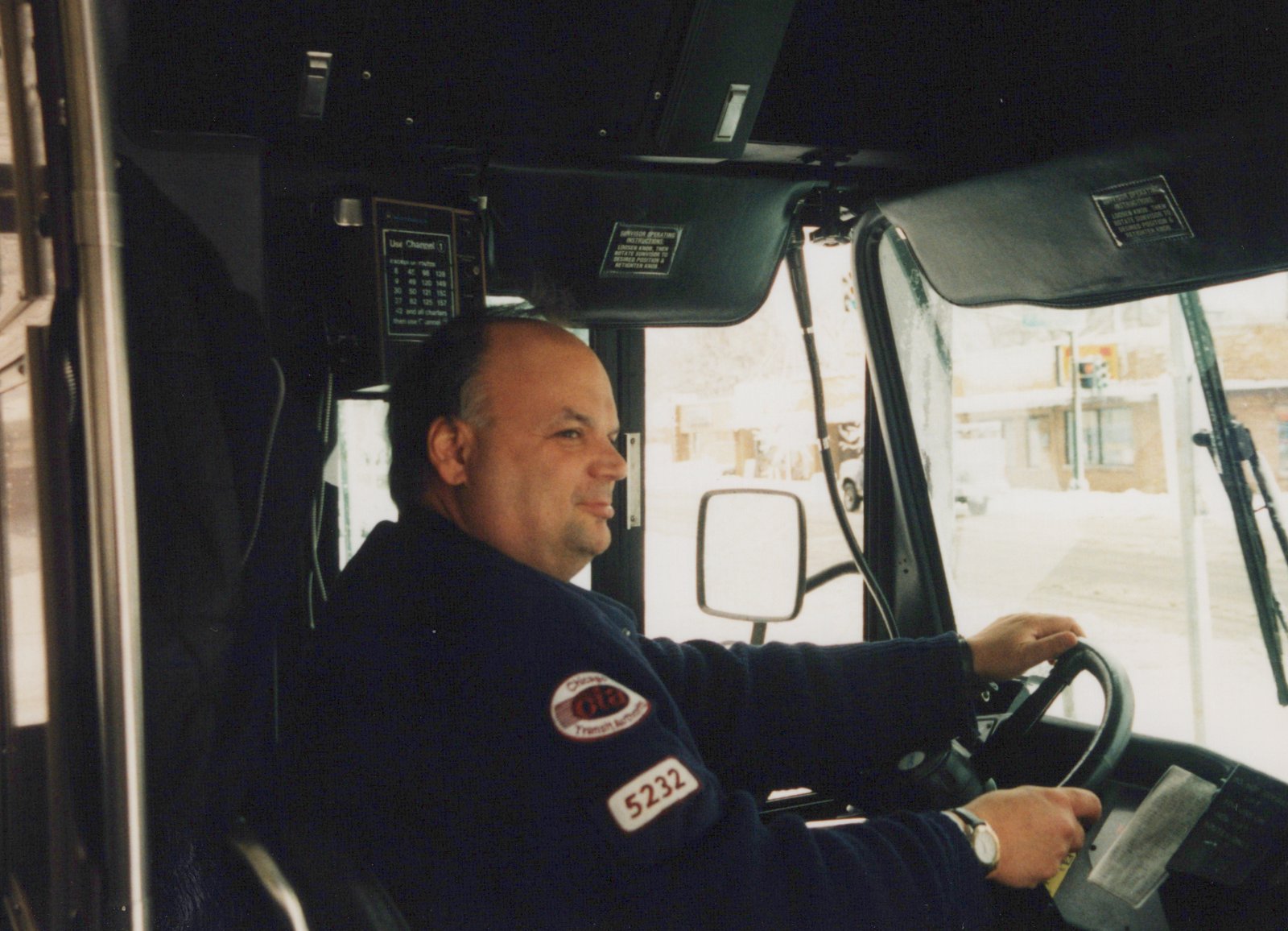 Interior view of a CTA bus driver at the wheel, wearing a dark blue uniform with patch and fleet number 5232, driving through a snowy city street.