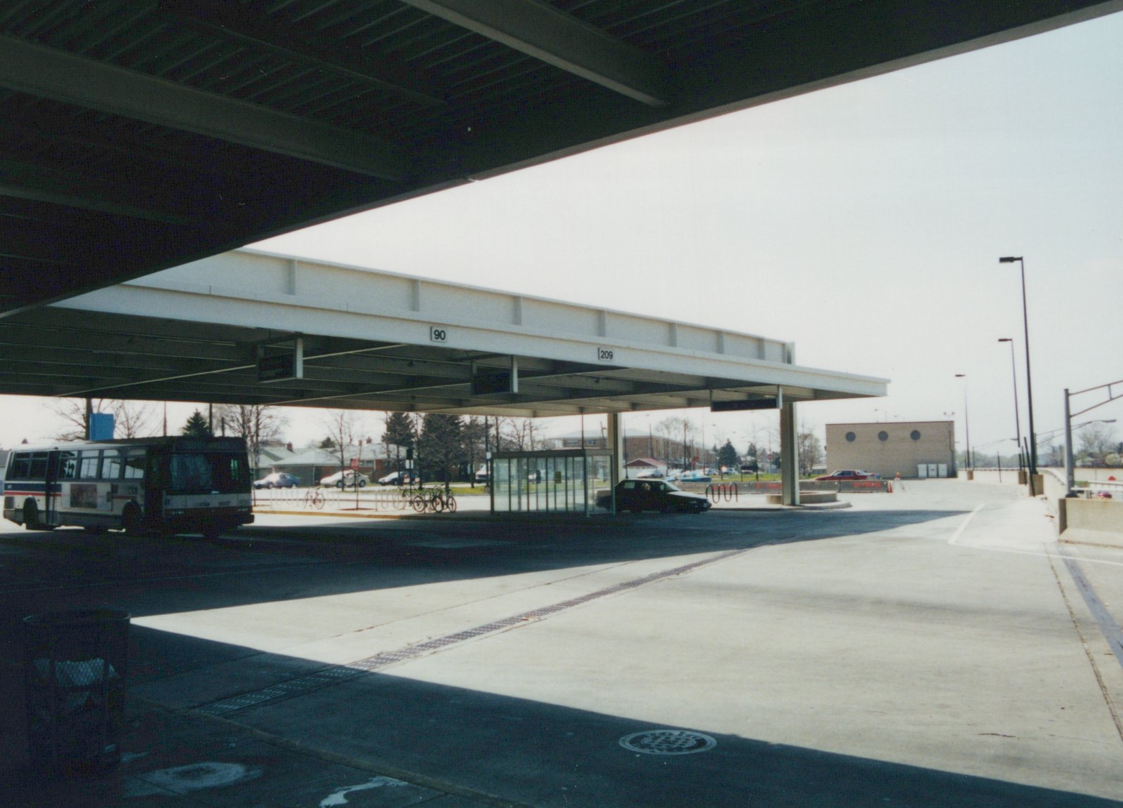 A white Pace bus with red and blue stripes is parked under a large, modern canopy at a bus terminal. The bus displays route 37 and fleet number 6007. Numbers 90 and 209 are visible on the shelter's beam.