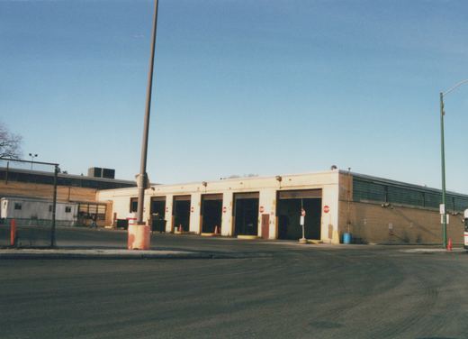 A long, single-story brick and block building with several large garage doors open, one person walking in the lot, and part of a white bus visible on the far right.