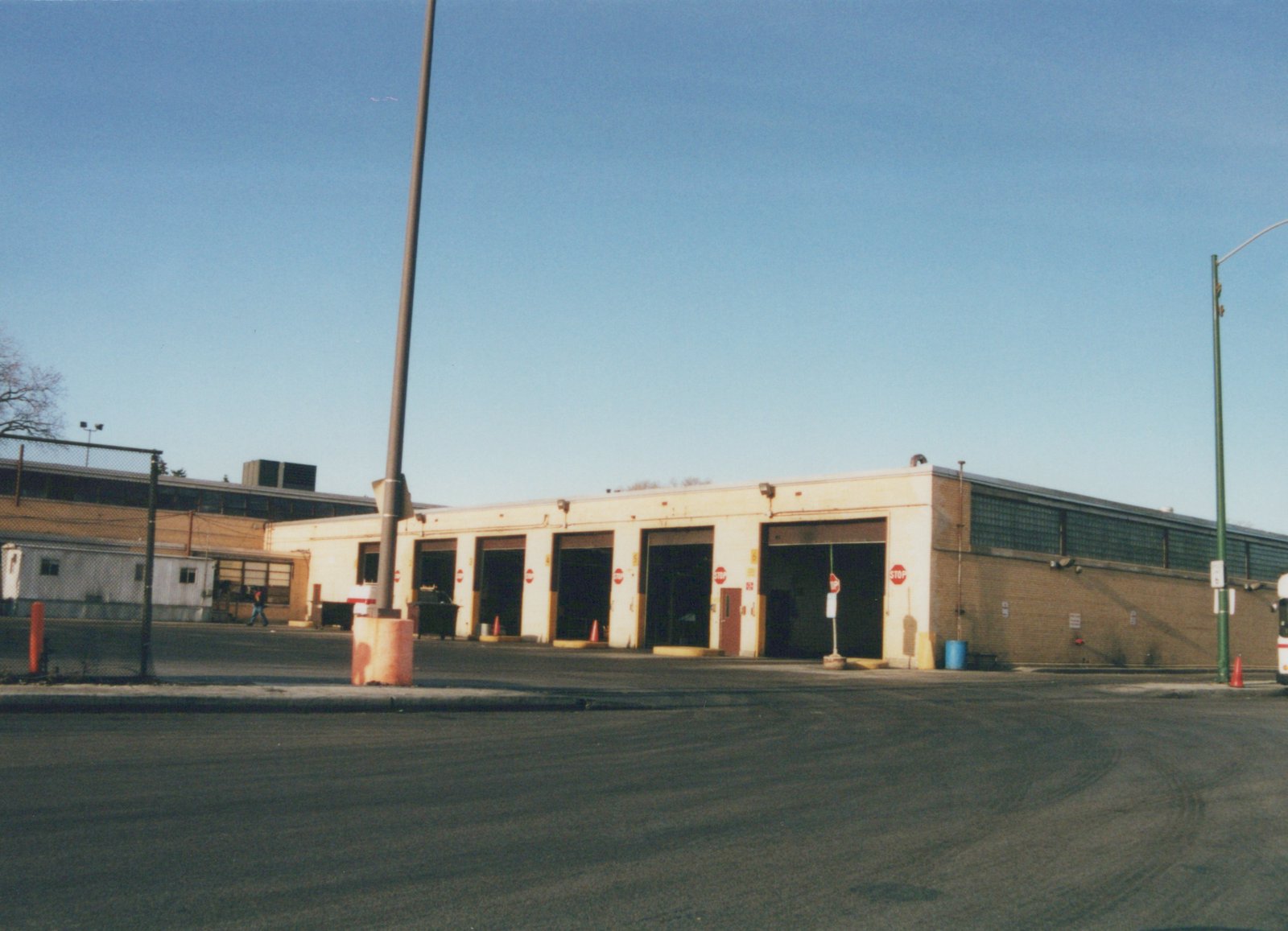 A long, single-story brick and block building with several large garage doors open, one person walking in the lot, and part of a white bus visible on the far right.