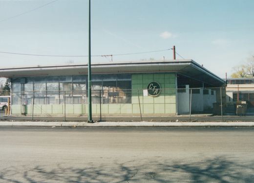 A street-level view of the Chicago Transit Authority's Forest Glen Garage, a mid-century modern building with a green tile exterior and a large circular CTA logo, with a bus visible inside through the windows.