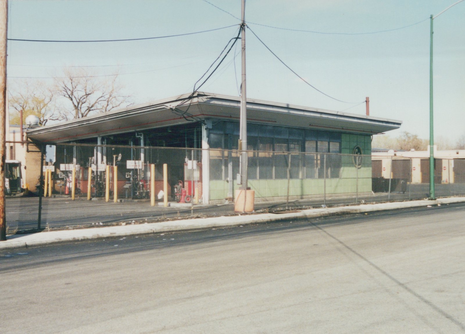 A low-angle view across a street of a long, single-story CTA garage building with a green tiled front and a flat roof, surrounded by a chain-link fence and utility poles under a clear sky.