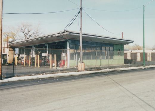 A low-angle view across a street of a long, single-story CTA garage building with a green tiled front and a flat roof, surrounded by a chain-link fence and utility poles under a clear sky.