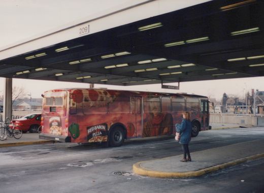 A red and orange bus with a full Red Baron Pizza wrap is parked under a covered bus stop on a winter day, while a woman in a blue jacket stands on the sidewalk.