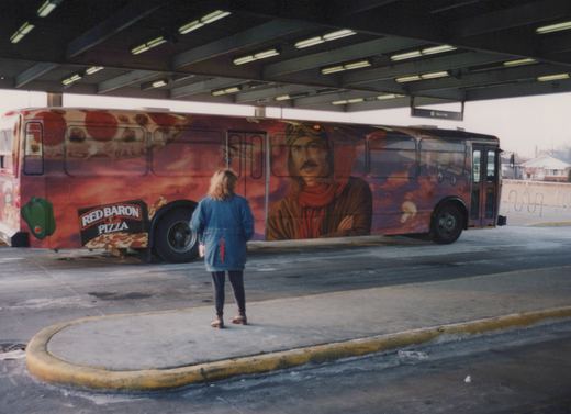 A side view of a Pace motor bus covered in Red Baron Pizza advertising, parked under a covered terminal structure with a person standing on the sidewalk.