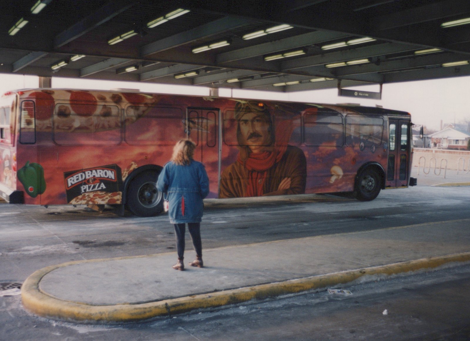 A side view of a Pace motor bus covered in Red Baron Pizza advertising, parked under a covered terminal structure with a person standing on the sidewalk.