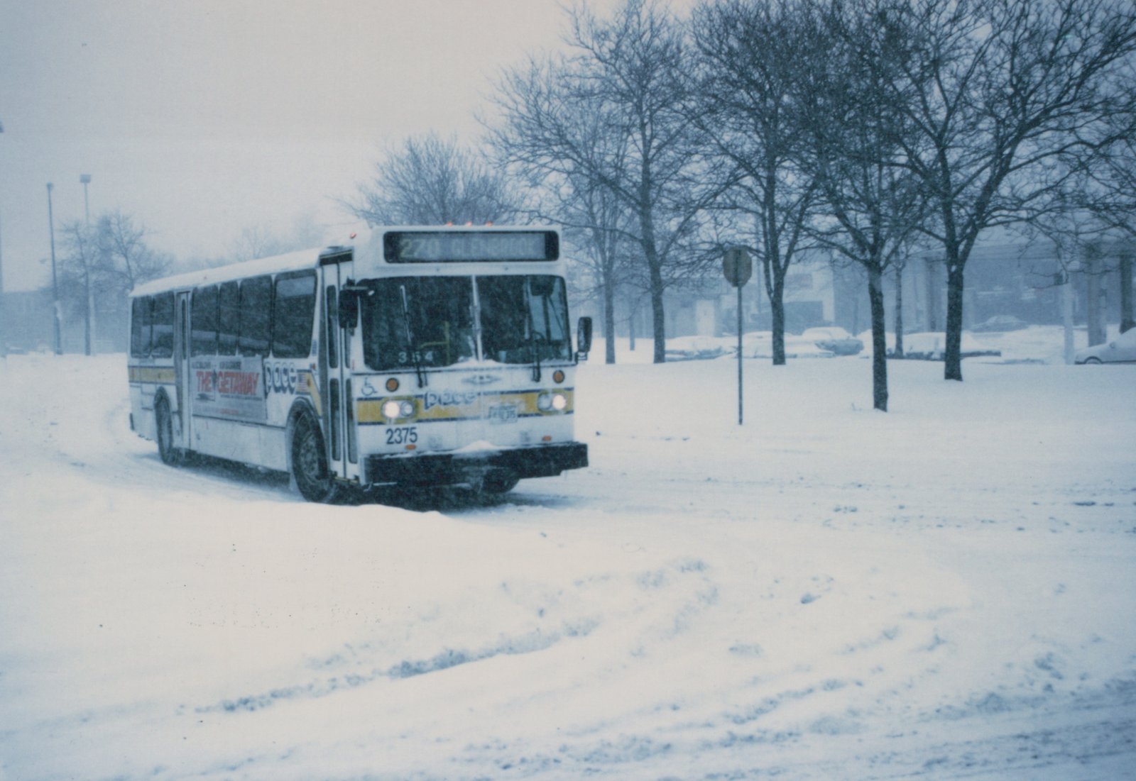 A white Pace bus is driving down a street covered in snow. The bus has a yellow stripe and displays route 270 to Glenbrook on its front sign. Bare trees line the street in the background.