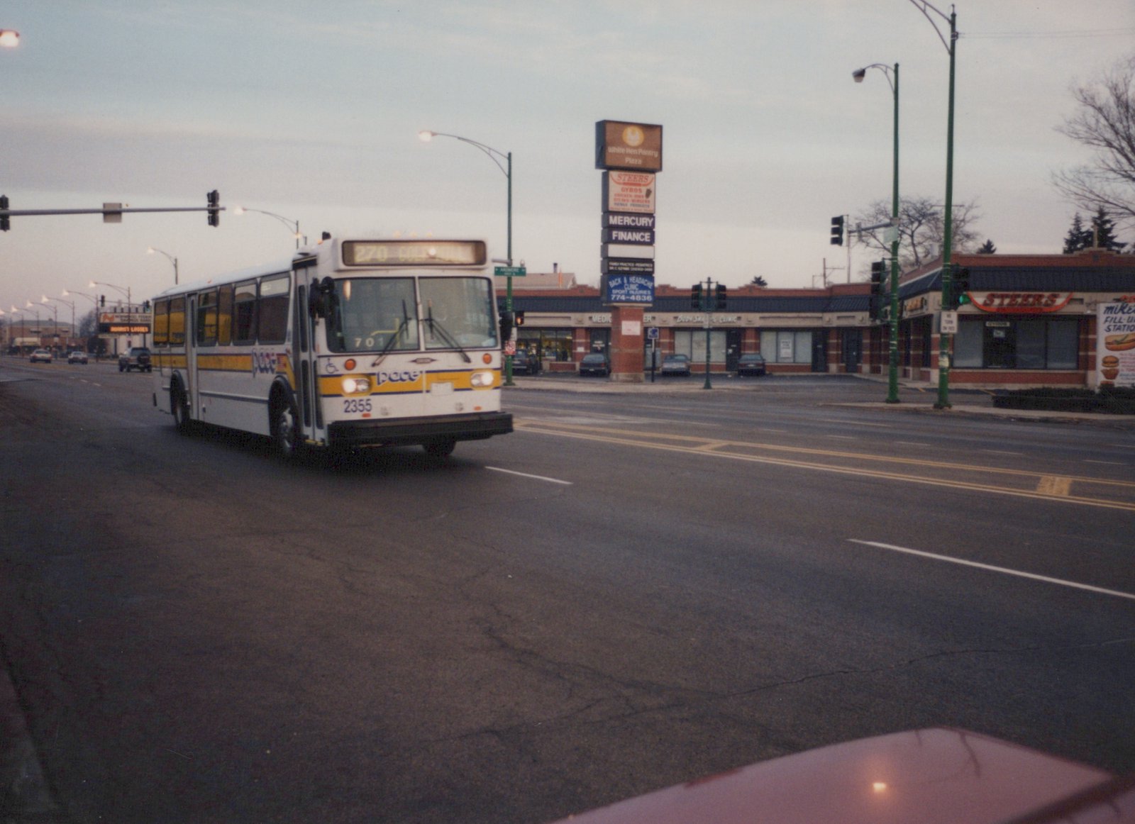 A white and yellow Pace bus with number 2355 is driving down a street at dusk with stores and a traffic light in the background.